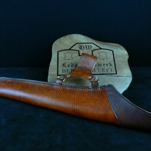 May include: A handcrafted leather quiver in shades of brown, with a dark brown accent at the tip. The quiver has a curved design and is held together with metal hardware. A wooden plaque with text is in the background.