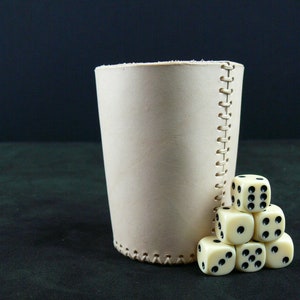 Puede incluir: Un cubilete de dados de cuero color beige claro con costuras, junto a una pila de seis dados de marfil con puntos negros. Los dados están dispuestos en forma de pirámide, con el cubilete a la izquierda.