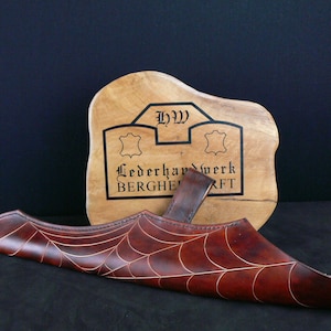May include: A dark brown leather sheath with a spiderweb design and gold stitching. The sheath is displayed in front of a wooden sign with the text "Lederwerk Bergheft".
