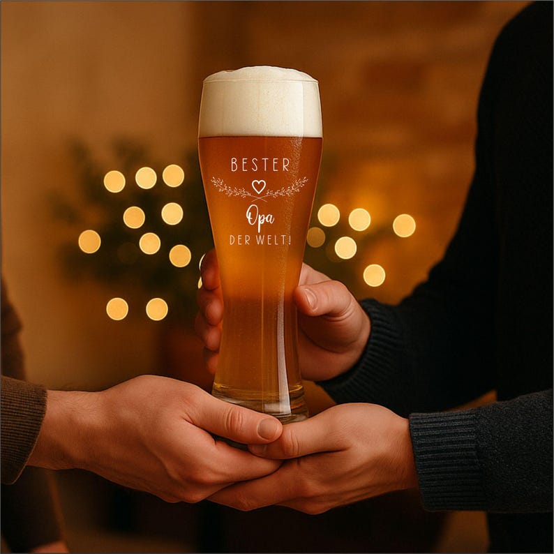May include: A tall beer glass filled with beer and foam, held by two hands. The glass has white text that reads "Bester Opa der Welt!" with a heart and floral design. The background is blurred with warm-toned lights.