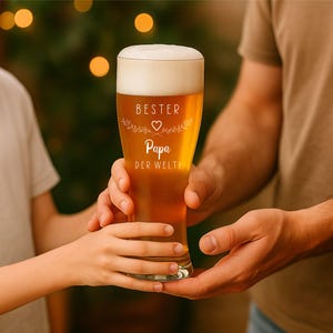 May include: A tall beer glass filled with golden beer and a thick white head. The glass has the words "BESTER Papa DER WELTI" in a decorative font. The glass is being held by two people.