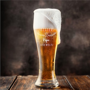 May include: A tall beer glass filled with golden beer and a thick, foamy head. The glass has the words "BESTER Papa DER WELTI" etched on it. The glass is on a wooden surface.