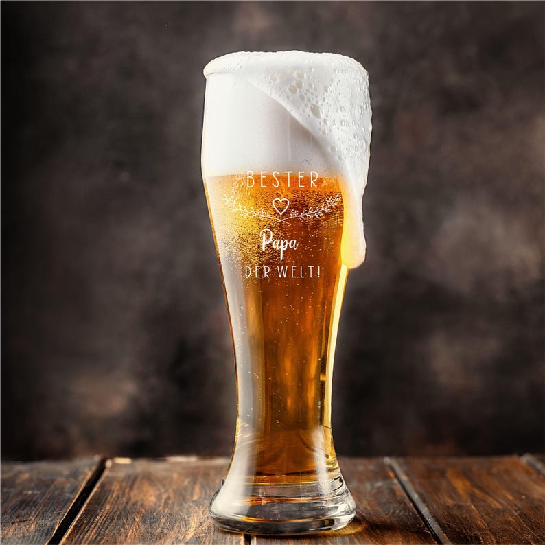 May include: A tall beer glass filled with golden beer and a thick, foamy head. The glass has the words "BESTER Papa DER WELTI" etched on it. The glass is on a wooden surface.