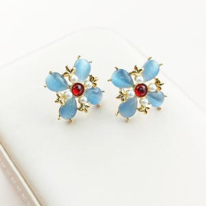May include: A pair of flower-shaped stud earrings. Each earring features four light blue teardrop-shaped stones, a central red stone, and small white pearls. The earrings have gold-colored metal accents.