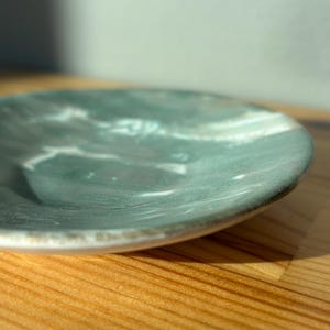 Puede incluir: Un plato de cerámica pequeño y redondo con un diseño veteado en verde azulado y blanco. El plato tiene un borde ligeramente elevado y está sobre una superficie de madera. La superficie del plato tiene un acabado brillante.