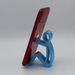 May include: A blue 3D printed phone stand shaped like a stick figure holding a red iPhone.
