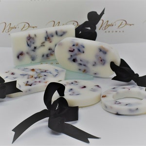 May include: Five white wax melts with dried flowers embedded in them. The melts are in various shapes, including a rectangle, oval, circle, and a ring.  Each melt has a black ribbon tied around it with a bow.