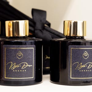 May include: Two black glass bottles with gold-coloured caps. Each bottle has a black label with gold lettering that reads "Night's Dream Aromas". A black ribbon is visible in the background.