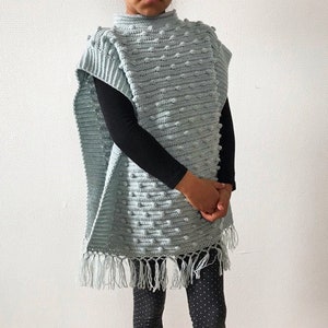 Könnte beinhalten: Ein hellblaues Häkelponcho mit einer strukturierten Noppenmasche und Fransenbesatz. Das Poncho hat einen hohen Kragen und wird von einem Kind getragen.