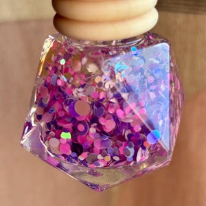 May include: A clear, faceted bottle filled with pink, purple, and iridescent glitter. The bottle has a light wood-coloured cap. The glitter is a mix of round and irregular shapes, creating a sparkling effect. The bottle is held up against a neutral background.