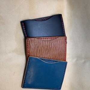 May include: Three leather cardholders in black, brown, and blue, stacked diagonally. Each cardholder has a contrasting stitch color: red on the black and brown, and black on the blue. The brown cardholder has a textured surface.