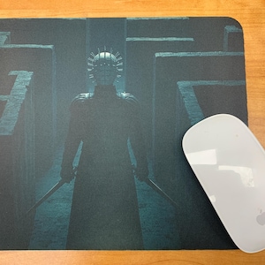 May include: A black and white mousepad with a dark blue and green image of a person wearing a mask and holding a weapon. The person is standing in a maze.