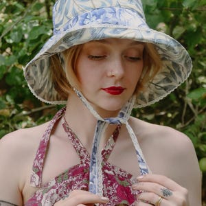 Puede incluir: Un sombrero de pescador con estampado de hojas en azul claro y crema, con un borde de encaje blanco en el ala y cintas para la barbilla. Se combina con un top halter color granate con estampado floral en blanco y verde y tirantes finos.