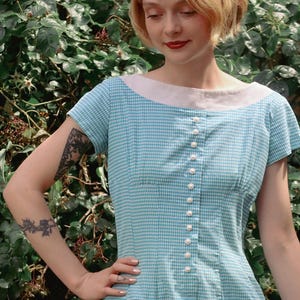May include: A light blue and white gingham dress featuring a wide, solid white boat neckline and short sleeves. The bodice is detailed with a vertical row of small, round, white pearl-like buttons running down the centre front of the garment.