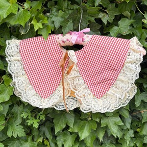 May include: A detachable collar featuring a red and white gingham pattern with wide cream-coloured lace trim along the edges. The collar is secured with thin brown ribbon ties at the centre front and is displayed on a padded floral fabric hanger.