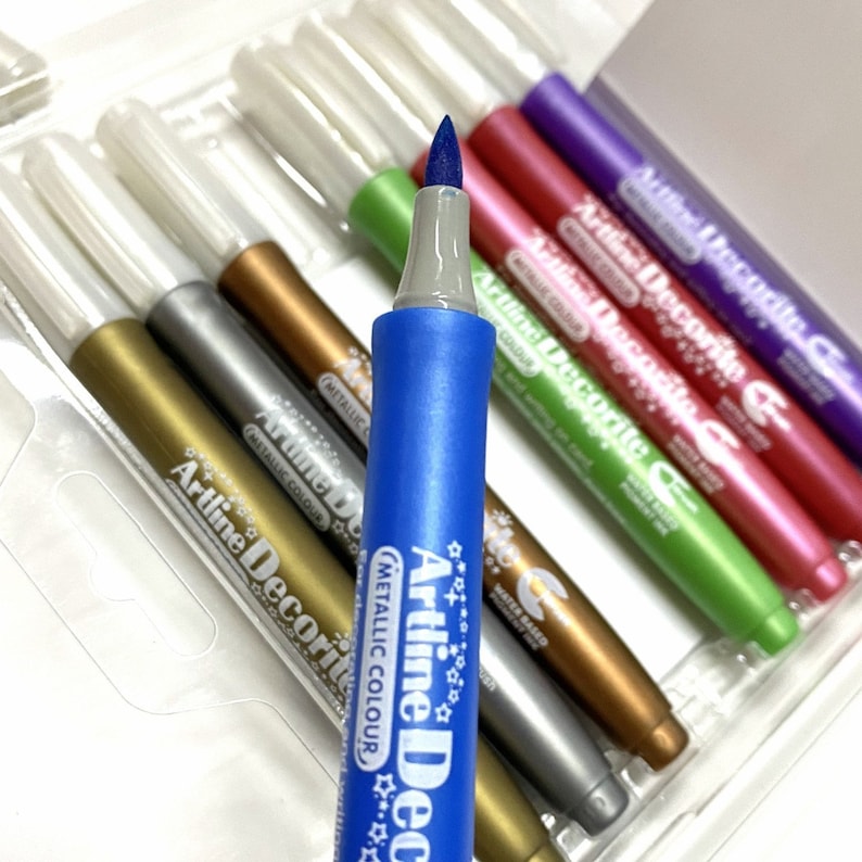 May include: A set of metallic markers with a blue marker in the foreground. The markers are labeled "Artline Decorite Metallic Colour" and have a star design. The markers are water-based.