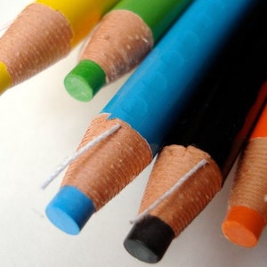 May include: Close-up of six sharpened colored pencils with exposed wood and white string wrapped around the ends. The pencils are blue, black, orange, green, and yellow.
