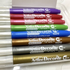 May include: A set of eight metallic paint markers with a brush tip. The markers are labeled "Artline Decorite" and "Metallic Colour". The markers are water-based pigment ink.