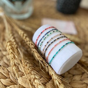 Peut inclure: Un ensemble de bracelets en perles colorées empilés sur un présentoir blanc. Les bracelets sont faits de petites perles colorées et sont disposés selon différents motifs.