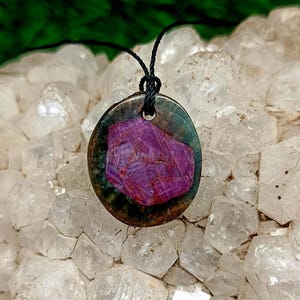 May include: Oval pendant featuring a vibrant, textured, deep magenta gemstone set in a dark metal frame. The pendant is suspended from a black cord. The gemstone has a crystalline appearance. The background is a light-colored, crystalline surface.