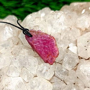 May include: A raw, ruby gemstone pendant with a black cord. The ruby is a deep pinkish-red color and has a rough, natural texture. The pendant is displayed on a bed of clear quartz crystals, with a green background.