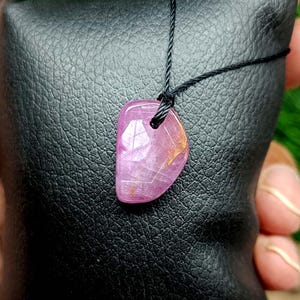 Peut inclure: Pendentif en pierre précieuse rose sur cordon noir. Le pendentif poli, de forme irrégulière, est translucide, avec des motifs internes et des inclusions dorées. Il repose sur une surface en cuir noir, avec un fond de feuillage vert.
