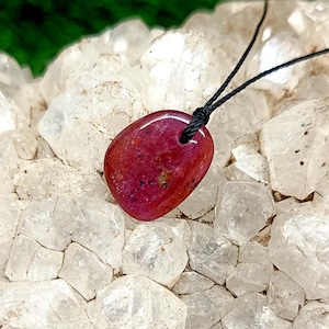 May include: A polished, oval-shaped ruby pendant on a black cord. The gemstone is a deep red color with subtle variations. The pendant is displayed against a backdrop of clear quartz crystals and a green background.
