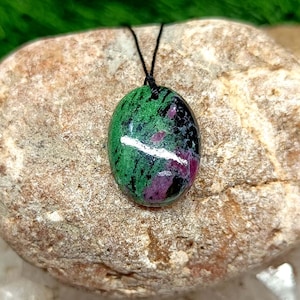 May include: Oval pendant with a black cord. The polished stone pendant displays a mix of green, black, and maroon hues. The stone is set against a natural, textured rock background.