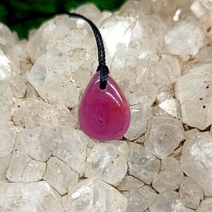 Pode incluir: Um pingente de rubi polido em forma de gota com um cordão preto. O pingente é de cor vermelho-rosada profunda e parece ser feito de uma pedra preciosa. O pingente é exibido em uma cama de cristais de quartzo transparentes.