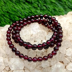 May include: A multi-strand bracelet composed of small, smooth, deep red garnet beads. The circular beads are uniform in size and strung together to form a continuous loop, showcasing a dark, translucent, and polished finish throughout the piece.