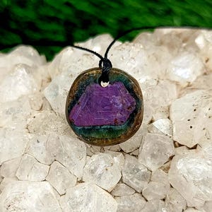 May include: A round pendant with a purple, geometric design set within a dark, iridescent border. The pendant is suspended from a black cord. The background features a cluster of clear, crystalline structures.