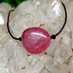 Peut inclure: Un pendentif en pierre précieuse rouge rubis poli, de forme ovale, monté sur un cordon noir. La pierre précieuse est le point central, sur un fond de formations cristallines claires. Le cordon est noué de chaque côté de la pierre.