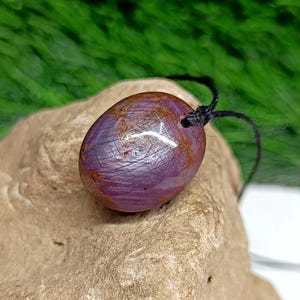 May include: A polished, oval-shaped pendant with a deep purple hue and reddish-brown accents. The gemstone is strung on a black cord. The pendant is resting on a light brown, textured surface, with a green background.