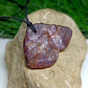 May include: A raw, irregularly shaped, deep burgundy gemstone pendant with a black cord. The stone has a rough, textured surface with lighter veins. The pendant is displayed on a light brown rock against a blurred green background.