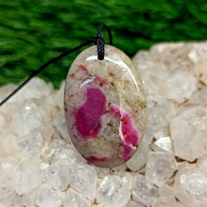 May include: Oval-shaped pendant with a black cord. The pendant features a light beige and gray stone with vibrant pink and red inclusions. The stone is polished and has a glossy finish. The pendant is set against a background of white crystals.