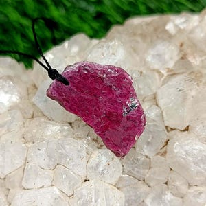 May include: A raw, deep red ruby gemstone pendant with a black cord. The ruby is irregularly shaped with a rough texture and sits on a bed of clear quartz crystals. The background features green foliage.