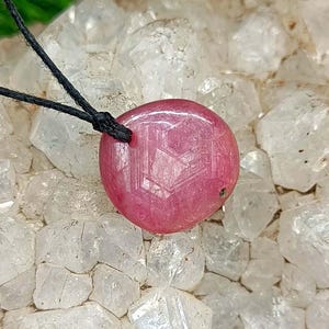 Peut inclure: Un pendentif en pierre précieuse rose, poli et rond, présentant un motif en étoile hexagonale visible à sa surface. La pierre est suspendue à un fin cordon noir, mettant en valeur une forme lisse et arrondie avec un effet d'étoile à six branches.