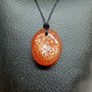 Op de afbeelding: Een ovaalvormige zonnesteen hanger met een levendige oranje kleur en interne gouden glitterinsluitingen. De edelsteen hangt aan een dun zwart koord en heeft een glad, gepolijst oppervlak dat het licht in het midden weerkaatst.