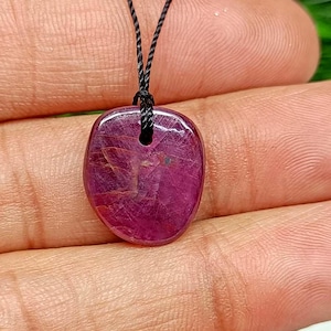 May include: A polished, oval ruby pendant on a black cord. The ruby is a deep, translucent red with internal veining. The pendant is suspended by a black cord, creating a simple and elegant necklace.