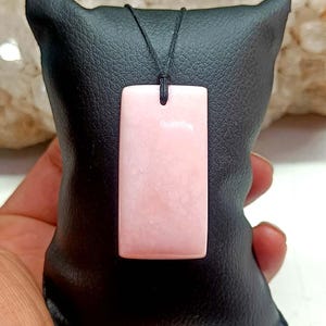 Könnte beinhalten: Ein rechteckiger Anhänger aus rosa Stein, der an einer dünnen schwarzen Kordel hängt. Der Anhänger hat eine glatte, polierte Oberfläche mit subtilen Variationen im hellrosa Farbton und zeigt eine klare Form, die vertikal an der Kordel hängt.