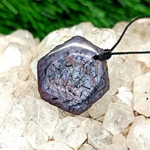 Peut inclure: Pendentif hexagonal avec un motif violet foncé et noir, suspendu à un cordon noir. Le pendentif est posé sur un fond de cristaux blancs et de feuillage vert.