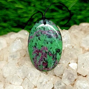 Può includere: Un ciondolo di forma ovale realizzato in zoisite verde con inclusioni di rubino magenta e macchie minerali nere. La pietra lucida presenta un piccolo foro nella parte superiore per un cordino nero, mostrando una superficie liscia e brillante.