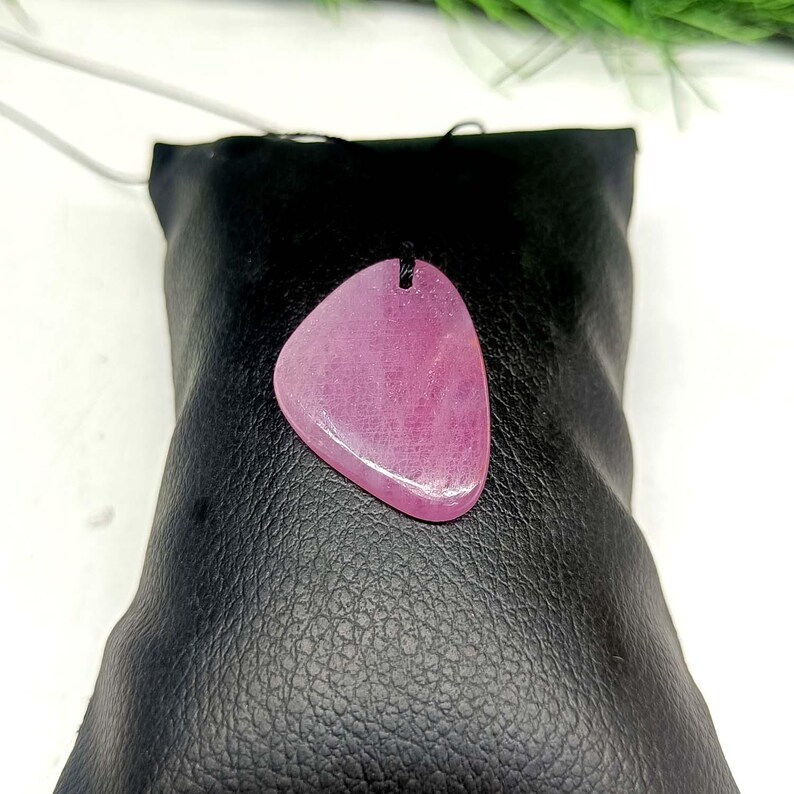 May include: A polished, teardrop-shaped pink gemstone pendant suspended on a thin black cord. The stone features a smooth, glossy surface with subtle internal variations in color and texture, centered on a black display cushion.