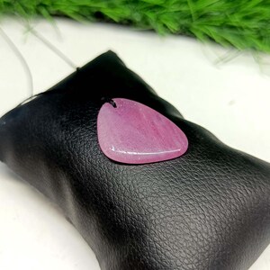 May include: A teardrop-shaped pink gemstone pendant with a smooth, polished surface and a small hole at the top for suspension. The pendant is resting on a black, textured, rectangular leather-like pouch with visible stitching.