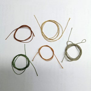 Pode incluir: Cinco rolos de barbante colorido em tons de marrom, dourado, verde e laranja, dispostos sobre uma superf&iacute;cie branca. Cada rolo &eacute; enrolado em forma circular, com as pontas do barbante estendendo-se para fora. O barbante parece ser feito de fibra natural.