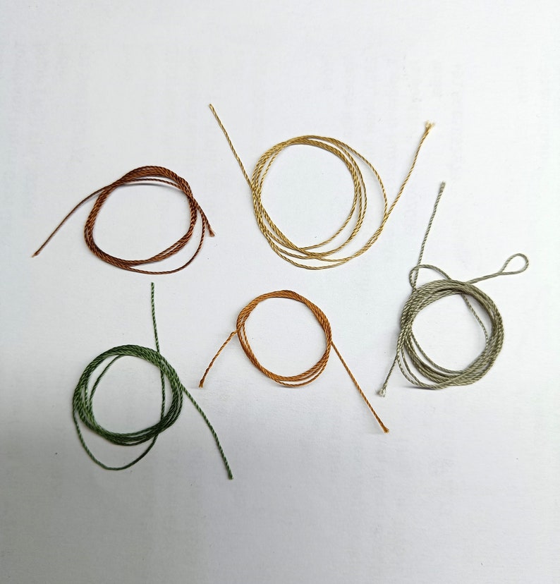 Pode incluir: Cinco rolos de barbante colorido em tons de marrom, dourado, verde e laranja, dispostos sobre uma superf&iacute;cie branca. Cada rolo &eacute; enrolado em forma circular, com as pontas do barbante estendendo-se para fora. O barbante parece ser feito de fibra natural.