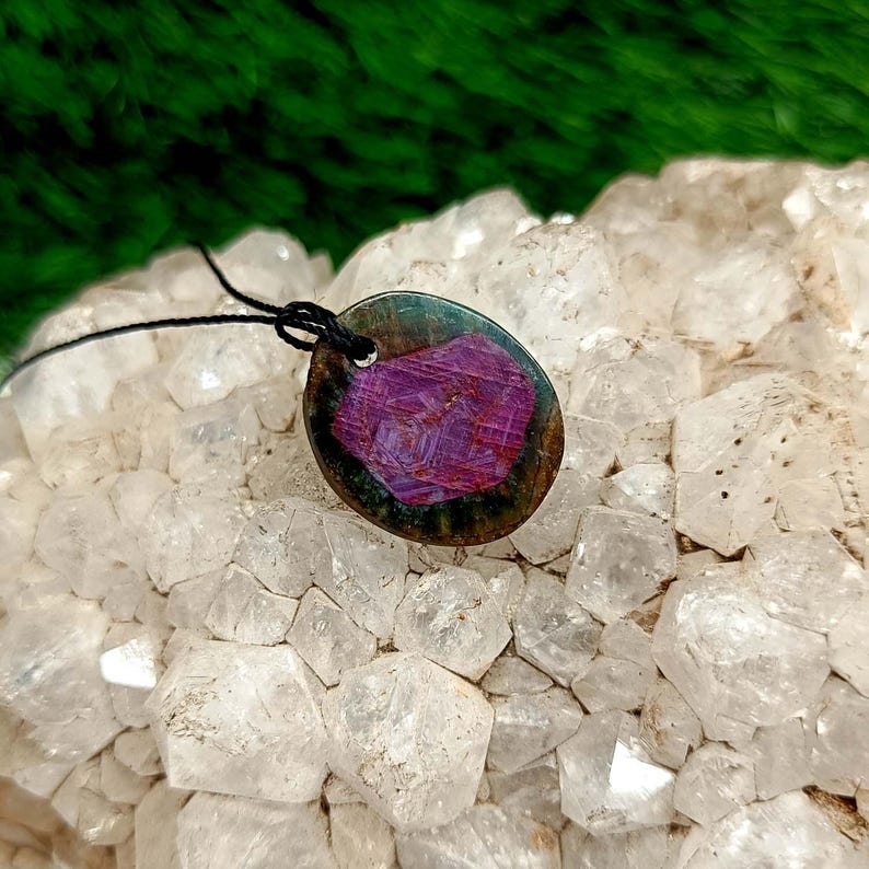 Peut inclure: Pendentif ovale orn&eacute; d'une pierre pr&eacute;cieuse violette vibrante, sertie dans une r&eacute;sine sombre et iris&eacute;e. Le pendentif est suspendu &agrave; un cordon noir. La pierre pr&eacute;cieuse pr&eacute;sente une structure cristalline. Le fond est un amas de cristaux de quartz clairs.