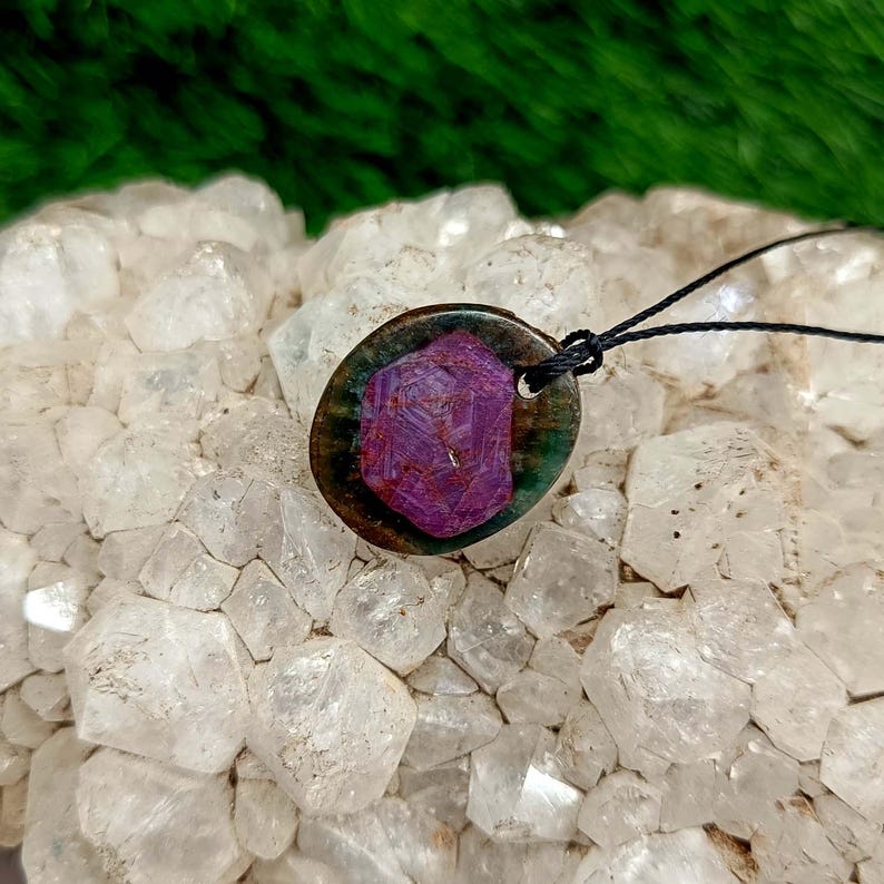 Peut inclure: Un collier pendentif avec une pierre pr&eacute;cieuse violet fonc&eacute; &agrave; facettes, sertie dans un cadre ovale fonc&eacute;. La pierre pr&eacute;cieuse est le point central, avec un cordon noir. Le pendentif repose sur un lit de pierres cristallines claires.