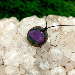 Pode incluir: Um colar com pingente redondo com uma vibrante pedra roxa embutida em um metal cor de bronze. A pedra tem uma apar&ecirc;ncia texturizada &uacute;nica. O colar pende de um cord&atilde;o preto, criando um contraste marcante.