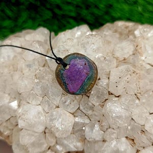 Pode incluir: Um pingente redondo com um centro roxo e iridescente, embutido em um metal de cor bronze. O pingente pende de um cord&atilde;o preto. O fundo &eacute; um aglomerado de cristais brancos, com folhagem verde vis&iacute;vel no canto superior esquerdo.